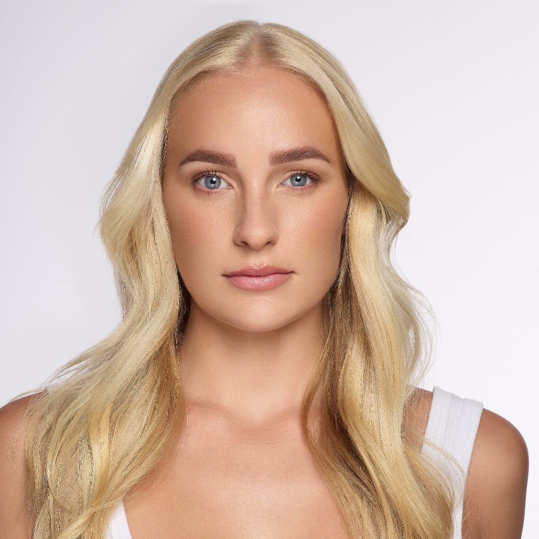 A close-up portrait of a blonde woman with long wavy hair, wearing a white top against a clean light gray background.