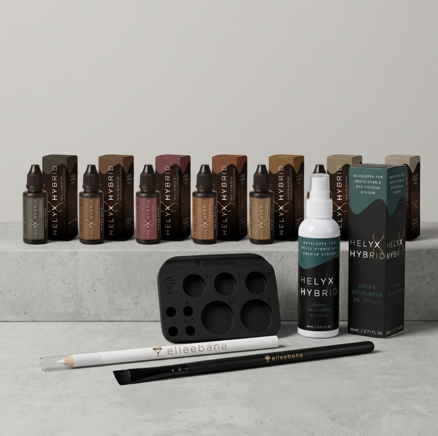 A Helyx Hybrid hair color kit displayed on a light gray stone surface against a neutral gray background. The kit includes multiple small bottles of brow stain in various shades, a white creme developer bottle, a black color mixingpalette, a white mapping pencil, and a black angled applicator brush, all branded with Helyx Hybrid and Ellebane.