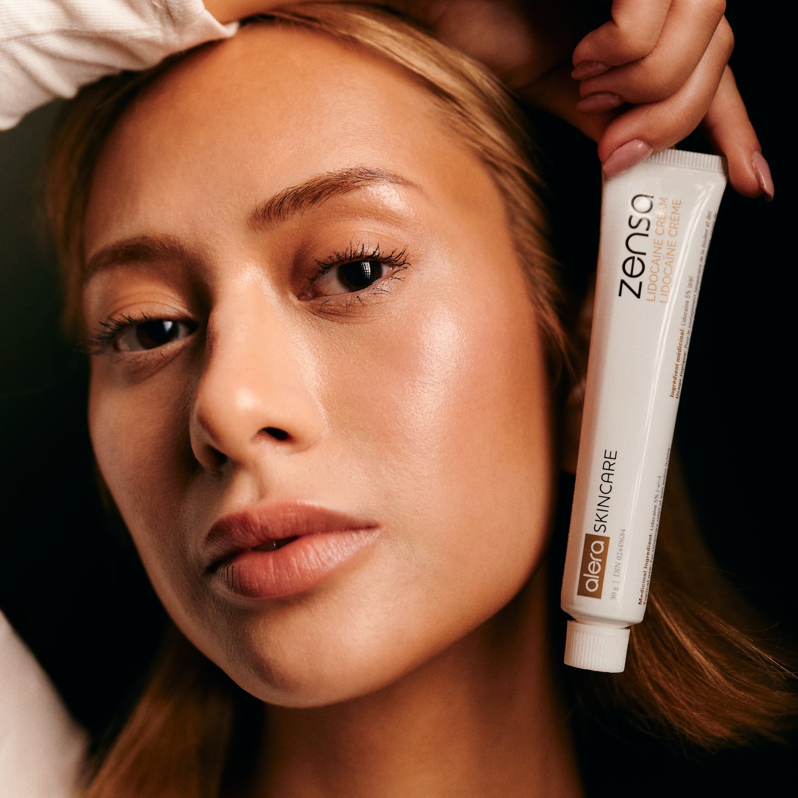 A close-up lifestyle shot of a woman holding a white tube of Zensa Numbing Cream against a dark background.
